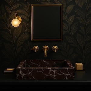 Op de afbeelding: Een donkerbruine marmeren badkamer wastafel met een gouden kraan en twee gouden handvatten. De wastafel is geplaatst tegen een zwarte muur met gouden bladbehang met een bladmotief. Een gouden spiegel hangt boven de wastafel.