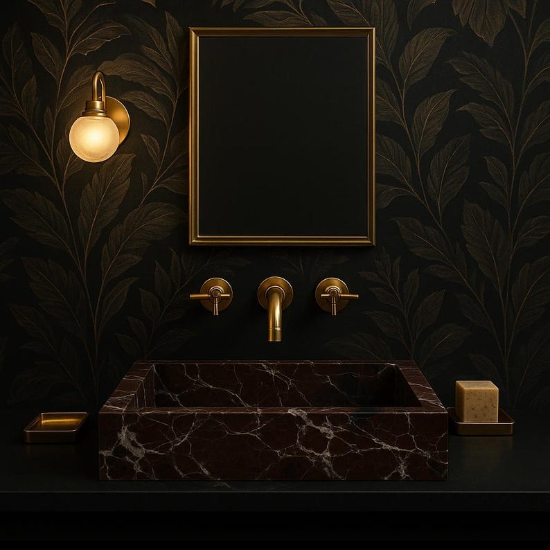 Op de afbeelding: Een donkerbruine marmeren badkamer wastafel met een gouden kraan en twee gouden handvatten. De wastafel is geplaatst tegen een zwarte muur met gouden bladbehang met een bladmotief. Een gouden spiegel hangt boven de wastafel.