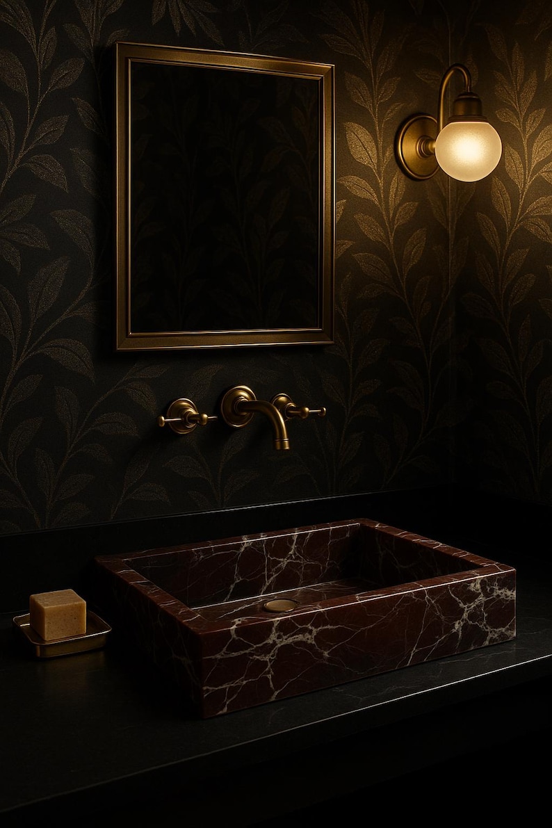 Op de afbeelding: Een donkerbruine marmeren wastafel met een gouden kraan en een zeepbakje. De wastafel is geplaatst tegen een zwarte muur met goudkleurig behang.