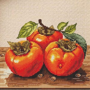 May include: A framed cross-stitch artwork featuring three vibrant orange persimmons with green leaves. The artwork is set against a light beige background, with a wooden frame. The image is surrounded by autumn-themed decorations, including leaves and cinnamon sticks.