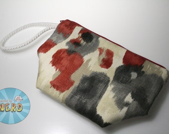 Pode incluir: Uma bolsa de zíper com padrão vermelho, cinza e bege com uma alça de corda branca. A bolsa tem um zíper vermelho e é feita de um tecido macio.
