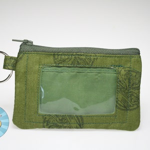 May include: Green fabric ID holder with a clear plastic window and a metal key ring. The holder has a zipper closure and a decorative pattern. The ID holder is approximately 5 inches wide and 3 inches tall.