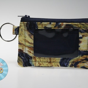 May include: A small, rectangular wallet with a clear ID window. The wallet has a zip closure and a metal ring for keys. The fabric has a marbled pattern in yellow, brown, and blue.
