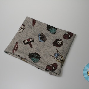 May include: A beige fabric square with a pattern of comic book characters and symbols. The characters include Spider-Man, Hulk, Thor, and Captain America. The fabric is folded in half and has a slight crease in the middle.