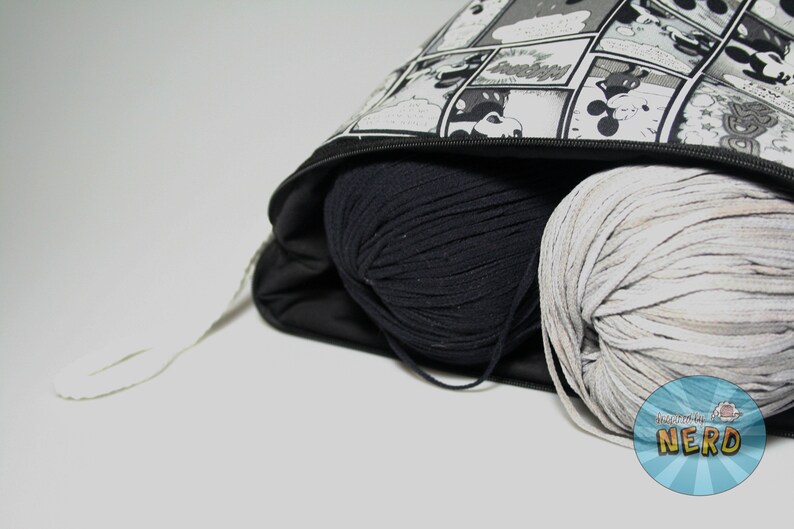 May include: A black and white comic book print zippered bag with a black drawstring. The bag is filled with two balls of yarn, one black and one white.