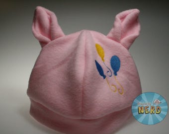 May include: A light pink fleece hat featuring two upright pointed ears at the top. The front of the hat is embroidered with a design of three balloons in yellow and blue colors, with swirling strings attached to each balloon.