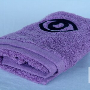 May include: A purple bath towel with a black embroidered eye design. The towel is folded in half and the eye design is centered on the towel.