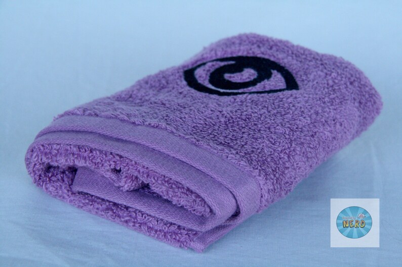 May include: A purple bath towel with a black embroidered eye design. The towel is folded in half and the eye design is centered on the towel.