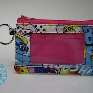 May include: A small, rectangular wallet with a clear ID window and a pink zipper. The wallet features a colorful cartoon print with a silver key ring attached.