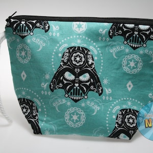May include: Teal zippered pouch featuring a black and white Darth Vader design. The design includes a stylized Darth Vader helmet and decorative patterns. The pouch has a white rope handle and a black zipper.