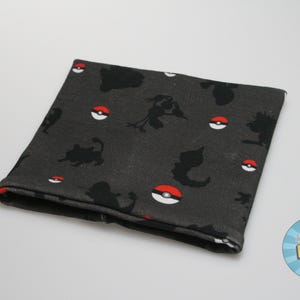 May include: A dark gray fabric neck gaiter featuring a pattern of black silhouettes of various creatures and red and white Poké Ball designs. The fabric is folded over, revealing a red edge.