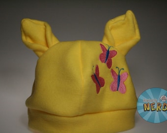 Op de afbeelding: Gele fleece-hoed met drie geborduurde vlinders in roze en blauw. De hoed heeft grote, slappe oren.