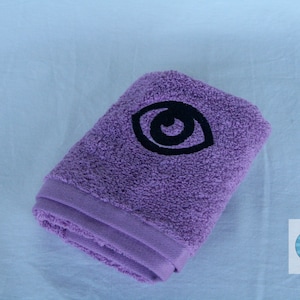 May include: A purple towel with a black embroidered eye design.