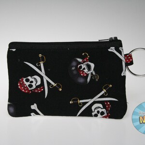 Puede incluir: Monedero negro con cremallera y estampado de calavera pirata y huesos cruzados. El diseño presenta calaveras blancas con pañuelos rojos y espadas cruzadas sobre un fondo negro. Incluye un anillo de metal plateado.