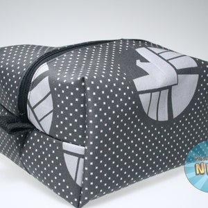 May include: A black and white zippered pouch with a white polka dot pattern and a gray logo. The logo is a stylized design of a bird with wings spread. The pouch is labeled "invented by NERD".