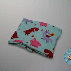May include: A folded, light blue fabric item with a repeating pattern of cartoon dinosaurs in red, pink, and purple. The dinosaurs are set against a background of small pink hearts. The item appears to be a neck warmer or similar accessory.