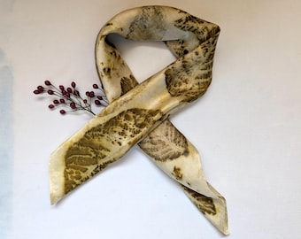 May include: A beige silk scarf with a leaf print in shades of brown and green. The scarf is tied in a knot and the ends are hanging down.