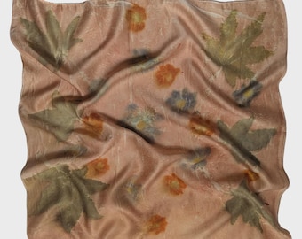 May include: A square, peach-colored scarf with a botanical print. The scarf features large, olive-green leaves and small, orange and blue floral patterns. The fabric is soft and draped, creating a textured appearance.