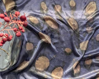 May include: A blue silk scarf with leaf imprints in shades of tan and brown. The scarf is draped, showing the texture of the fabric. A glass vase with red berries is in the foreground.