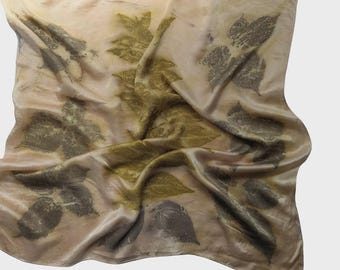 May include: A silk scarf with a botanical leaf print. The scarf displays a pattern of leaves in shades of green and grey, set against a beige and tan background. The fabric has a subtle sheen, and the scarf is draped.