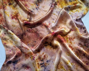 May include: A silk scarf with a hand-dyed pattern. The fabric displays a mix of colours, including pink, yellow, and brown, creating a unique, artistic design. The scarf's texture is smooth and lustrous, with visible folds and drapes. The scarf is made of silk.