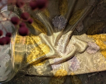 May include: A still life composition featuring a glass vase with dark red berries and a draped fabric. The fabric is a mix of gold, olive green, and gray, with floral and leaf patterns. The lighting creates shadows and highlights.