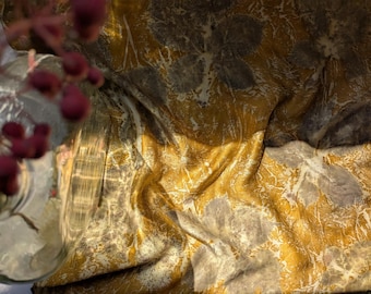 May include: Close-up of a fabric with a gold and gray marbled pattern. The fabric is draped, creating folds and shadows. The texture appears soft and luxurious. A glass vase with red berries is in the foreground.