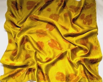 Può includere: Una sciarpa di seta giallo dorato con un motivo ripetuto di stampe di foglie arancioni. La sciarpa è drappeggiata, mostrando la lucentezza del tessuto. Le stampe delle foglie variano in dimensioni e sono sparse.