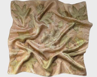 May include: A square, peach-coloured silk scarf with a botanical print. The scarf features a pattern of pressed leaves in shades of green and brown. The fabric has a soft, flowing drape.