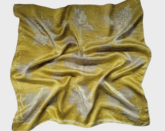 May include: Mustard yellow silk scarf with a subtle floral pattern in shades of beige and cream. The scarf is draped, showing the texture and sheen of the fabric. A stylish accessory.