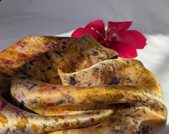 May include: A silk scarf with a botanical print in shades of gold, brown, and purple. A vibrant red flower rests on the white surface next to the scarf. The scarf is draped and folded, showcasing its intricate design.