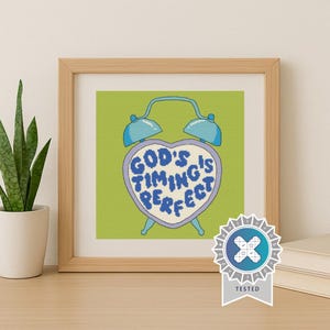 May include: A framed cross-stitch artwork featuring a light blue alarm clock shaped like a heart on a lime green background. The clock face contains the dark blue text "GOD'S TIMING IS PERFECT" in a pixelated font style, framed in light wood.