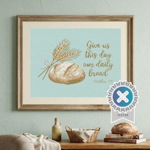 May include: Cross-stitch artwork in a wooden frame, depicting a loaf of bread and wheat. The text reads "Give us this day our daily bread" with the reference Matthew 6:11. The background is a pale blue.