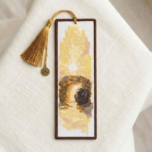 May include: A handmade bookmark with a cross-stitch design. The bookmark features a rectangular shape with a brown border and a detailed image in shades of yellow, brown, and white. It has a gold tassel and a small fish charm attached.