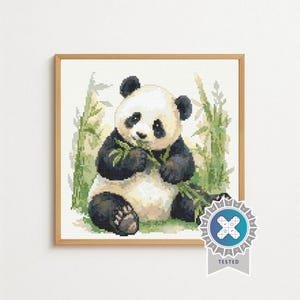 May include: A framed cross-stitch embroidery featuring a black and white panda sitting and eating green bamboo stalks. The panda is centered against a light background with green foliage. A blue and white circular badge with the word TESTED is visible.