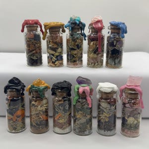 Peut inclure: Une collection de petits flacons en verre remplis d'herbes séchées et de cristaux. Chaque flacon est scellé avec un bouchon de liège et surmonté de cire colorée, notamment rouge, jaune, bleu et rose. Les flacons sont disposés en deux rangées.