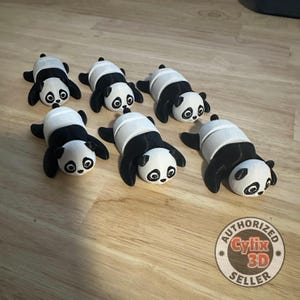 Peut inclure: Six figurines de panda noir et blanc couchées sur une surface en bois. Les pandas sont tous dans une pose similaire, la tête inclinée sur le côté et les yeux fermés. Les figurines sont en plastique blanc lisse et ont des marques noires autour des yeux, des oreilles et des pattes.