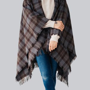 Könnte beinhalten: Ein karierter Poncho in Grau- und Brauntönen mit Fransen. Der Poncho wird über einem weißen Pullover und einer blauen Jeans getragen. Das Karomuster ist ein klassisches Design, geeignet für Herbst- und Wintermode.