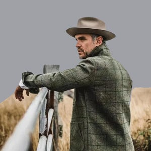 May include: A man wearing a grey fedora hat and a green plaid coat leans against a white fence. The coat has large pockets and a collar. The background is a field with tall grass.