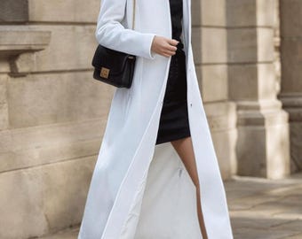 Puede incluir: Un abrigo largo blanco con cuello alto y un bolso negro con cadena dorada. El abrigo se combina con un vestido negro y botas marrones de tacón. El atuendo es adecuado para una ocasión formal o semiformal.