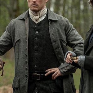 May include: A man in a gray wool coat, dark green vest, and white cravat. The coat has large pockets and button details. A black belt is visible. The background is blurred, suggesting an outdoor setting.