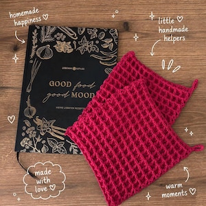 May include: A black hardback recipe book with gold botanical illustrations and the text "GOOD food good MOOD" alongside two square, magenta-coloured, waffle-textured crocheted potholders with small corner loops for hanging.