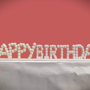 May include: A white "HAPPY BIRTHDAY" sign made of small, round, white objects. The sign is placed on a white surface against a red background. The letters are arranged in a horizontal line.