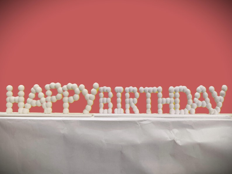 May include: A white "HAPPY BIRTHDAY" sign made of small, round, white objects. The sign is placed on a white surface against a red background. The letters are arranged in a horizontal line.