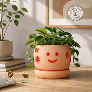 May include: A peach-colored planter with a smiling face design, red accents, and decorative flowers. The planter holds a green leafy plant. The text "TESTED & TRUSTED" and "MADE FOR MAKERS" are visible.