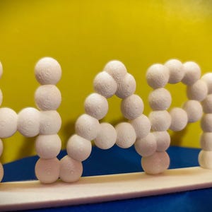 May include: A white 3D-printed sculpture spelling out the word "NAP". The letters are formed from stacked spheres with a textured surface. The background is yellow and blue.