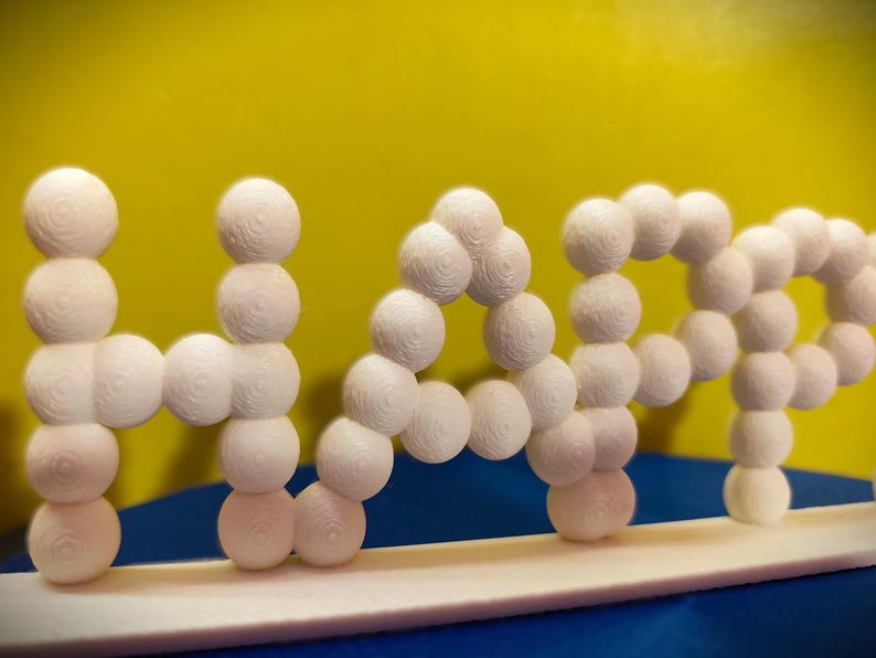 May include: A white 3D-printed sculpture spelling out the word "NAP". The letters are formed from stacked spheres with a textured surface. The background is yellow and blue.