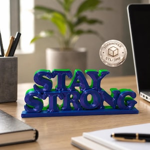 May include: A 3D-printed desk ornament with the words "STAY STRONG" in blue and green. The letters are layered and stand on a blue base. A 3DPrintFile logo is visible in the background. The ornament is on a wooden desk.