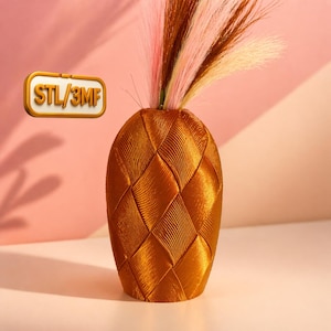 May include: A bronze-coloured vase with a woven texture, containing a bouquet of pink, brown, and cream ornamental grass. The vase is set against a pink and peach background. The text "STL/3MF" is in a gold rectangle.