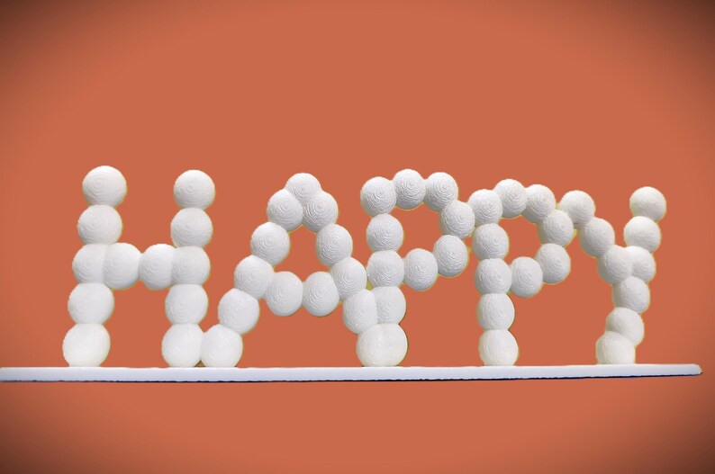 May include: White, textured spheres form the word "HAPPY" against an orange background. The letters are connected and rest on a white, horizontal base. The overall design is simple and cheerful, suitable for home decor.